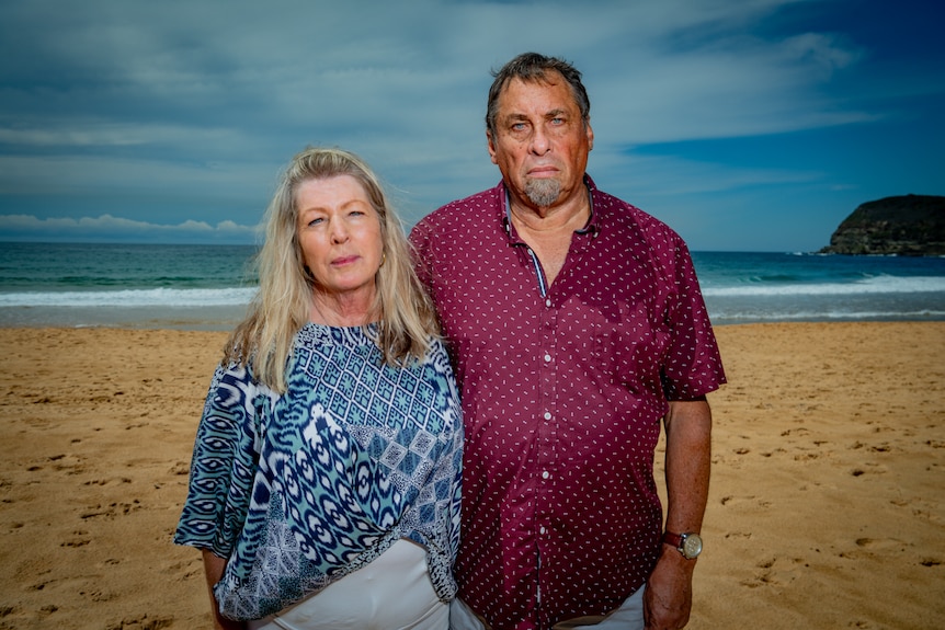 A man and a woman stand on a beach with their arms around each other. They have grave expressions on their faces.