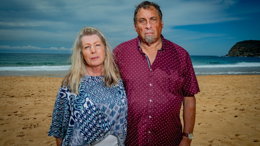 A man and a woman stand on a beach with their arms around each other. They have grave expressions on their faces.