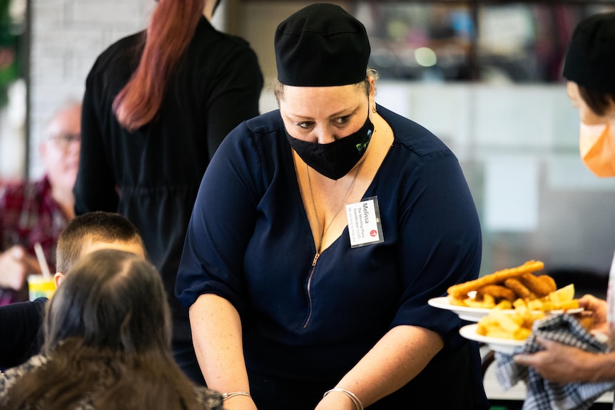 A woman in a mask serves customers at a table in a cafe.