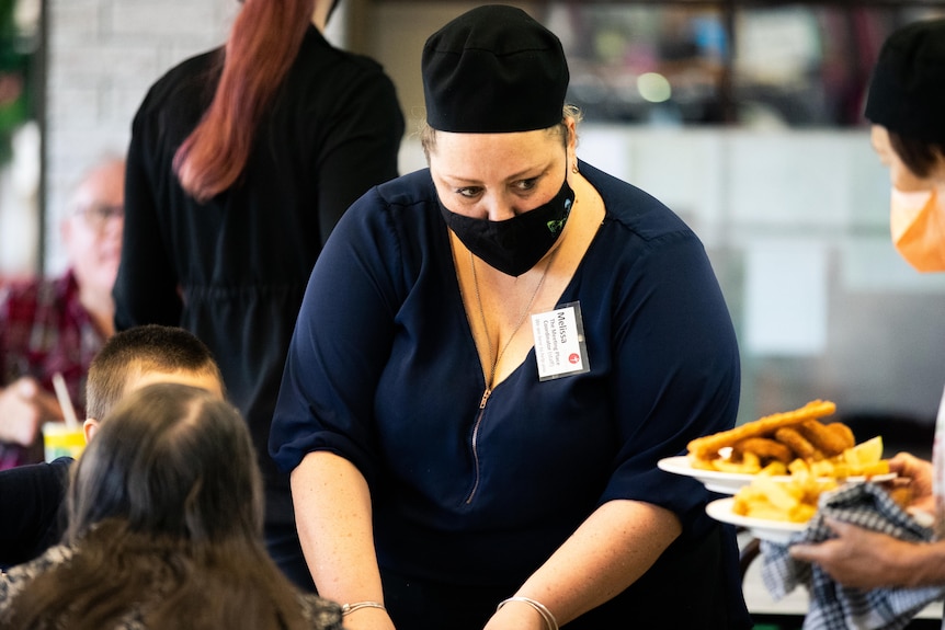 Une femme masquée sert des clients à une table dans un café.