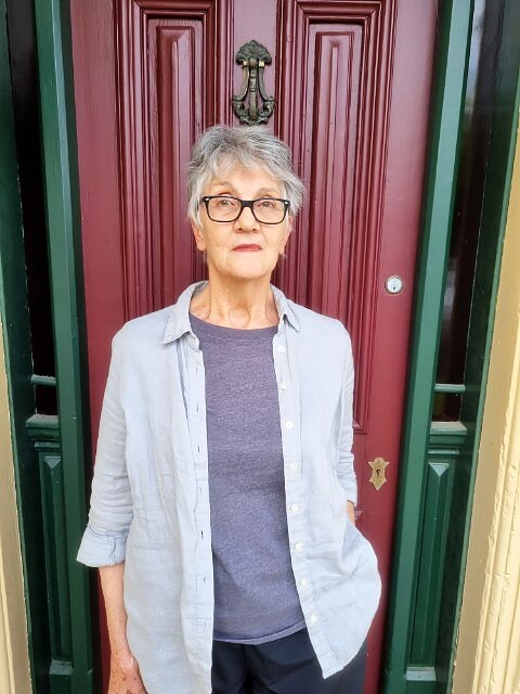 An older woman in glasses stands against a red door.
