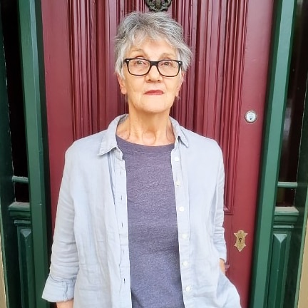 An older woman in glasses stands against a red door.