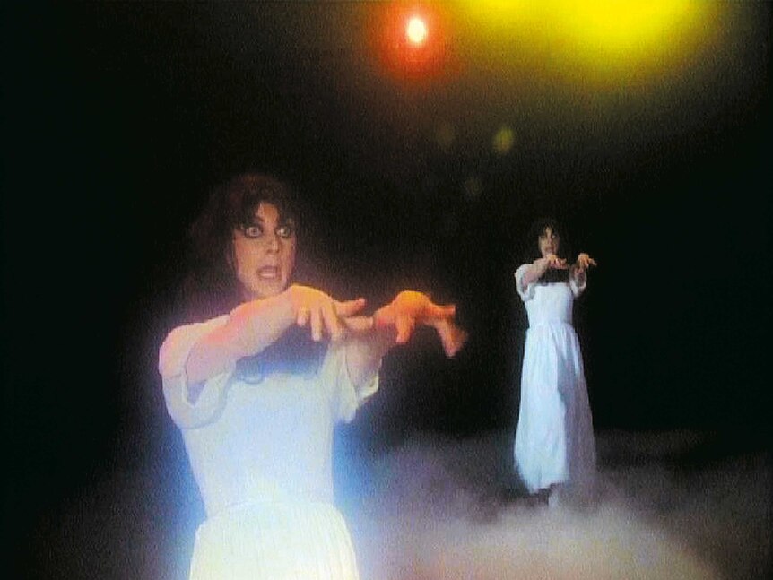 A man in drag holds his arm out in front of him, lights and smoke surround him