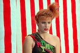 A woman in 80s dress stands in front of a red and white backdrop.