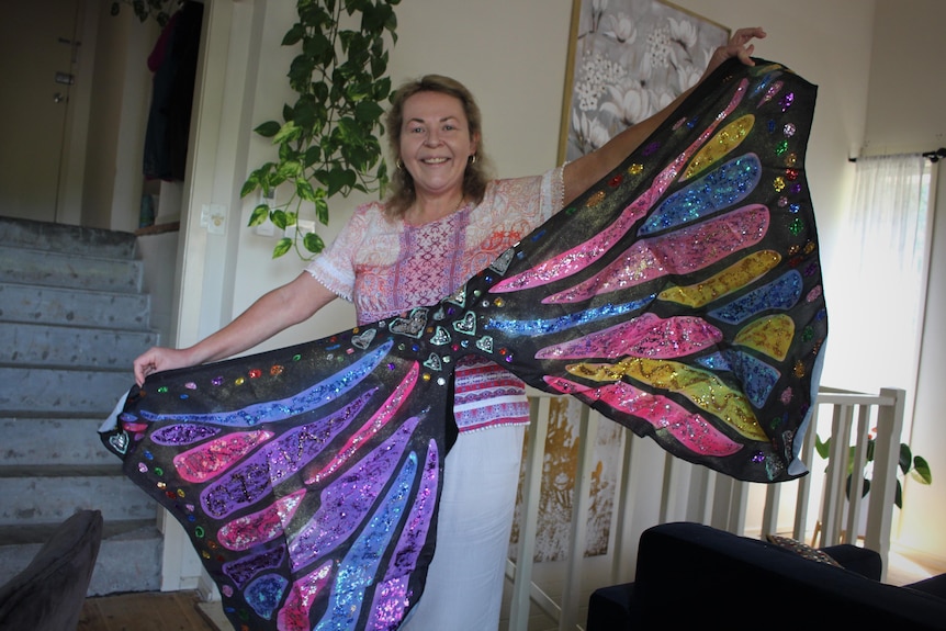 A middle aged woman, smiles holding open dress up painted butterfly wings. 