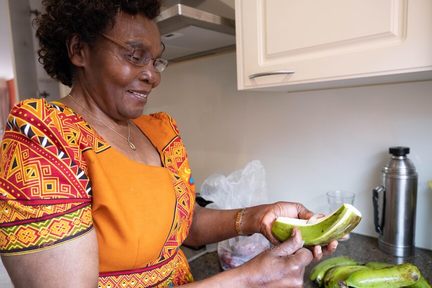 Theresa peels a plantain in her kitchen wearing an ornage dress and smiling. 