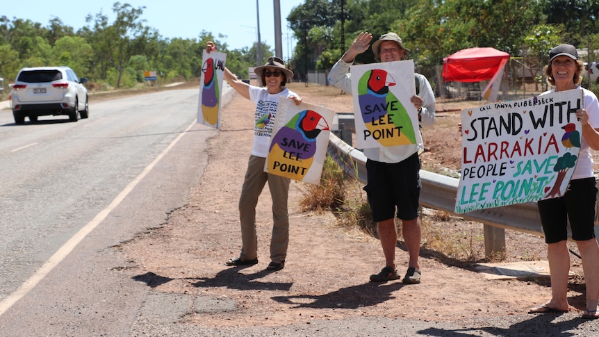 Three people stand by the side of the road holding 'save lee point' posters. They are happy.