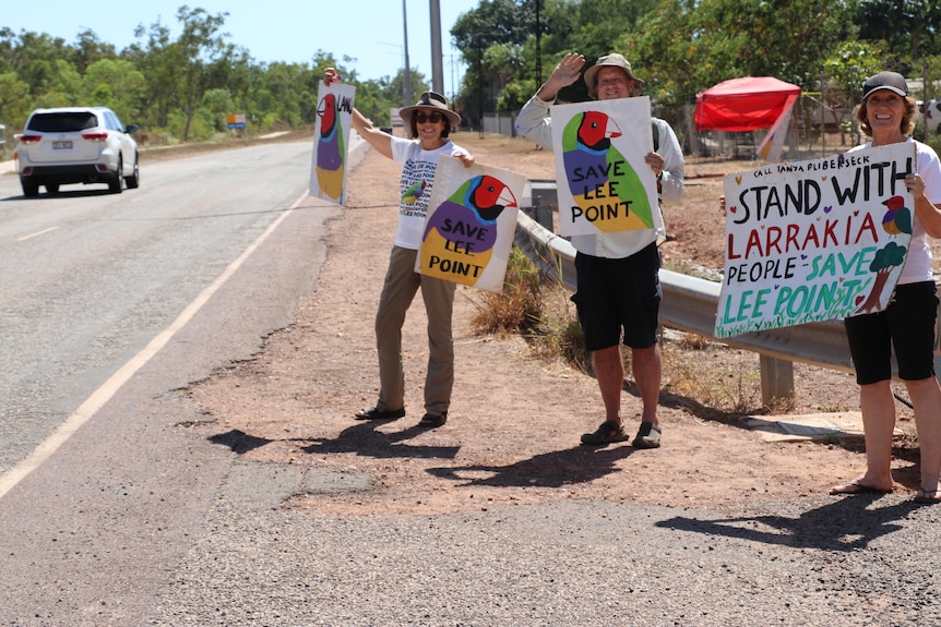 Three people stand by the side of the road holding 'save lee point' posters. They are happy.