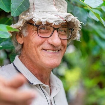 man iun spectacles and straw hat with hand extended outwars in front of leafy garden