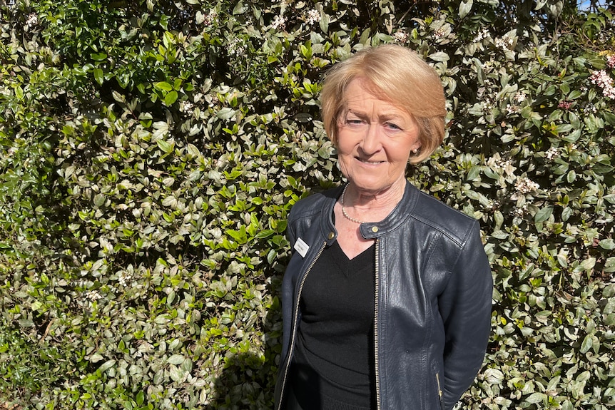 Christine wears a black top and leather jacket and has short blonde hair, she stands in front of a hedge