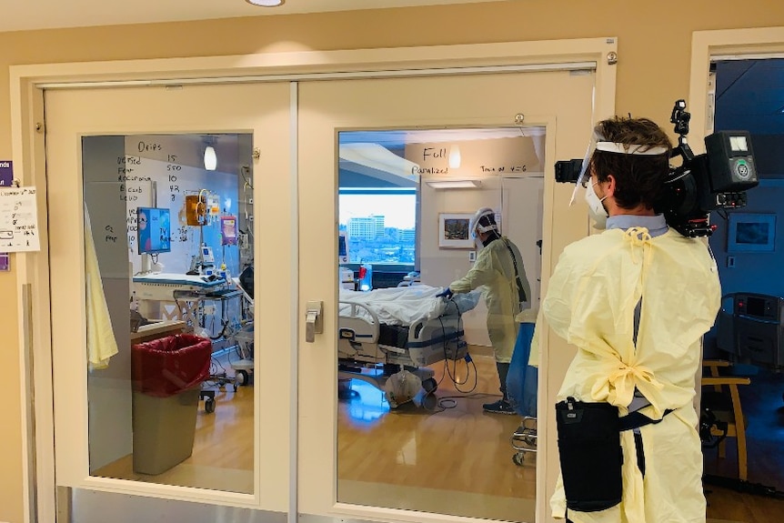 Cameraman in full PPE filming patient in hospital bed being cared for by nurse in full PPE.