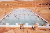 A sepia photograph of people swimming in a pool with red hills in the background.