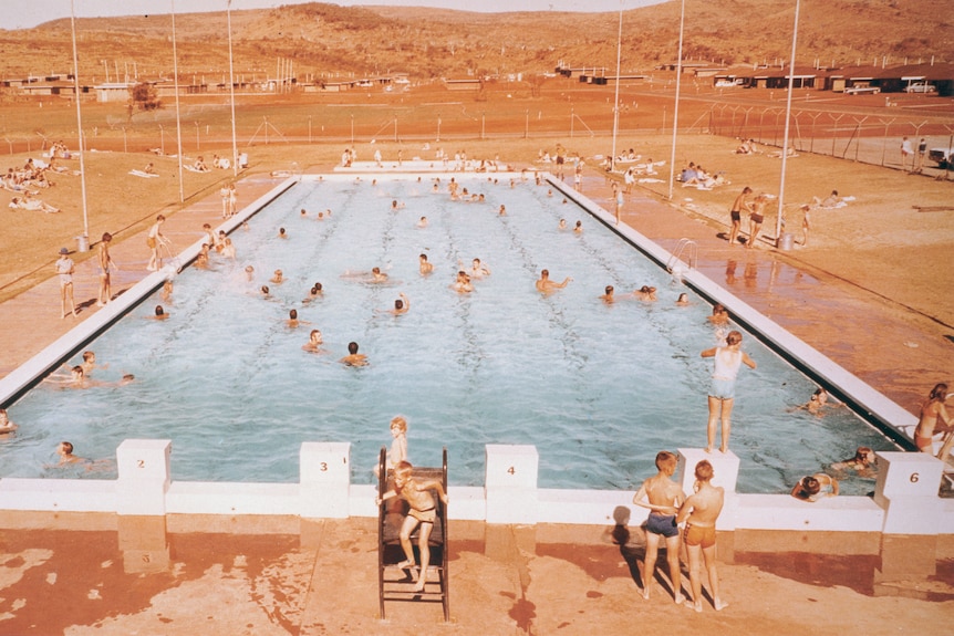 A sepia photograph of people swimming in a pool with red hills in the background.