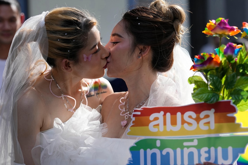 Two women in bridal dresses kiss in front of a poster to support gay marriage