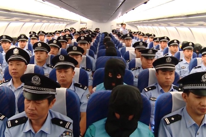 Chinese suspects on the plane. They are hooded and seated in the middle row and end rows, with police filling all other seats.