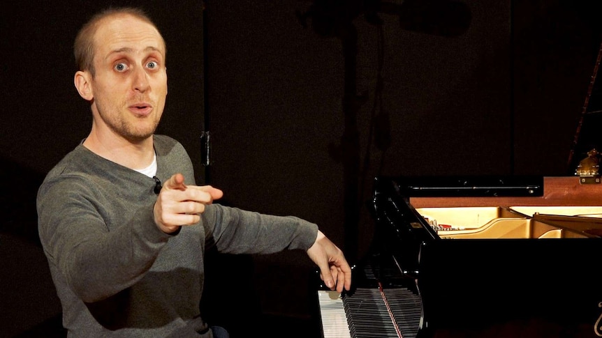 A man sitting at a piano points to the camera.