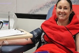 A woman wrapped in a red blanket sits in a blood donation chair smiling at the camera