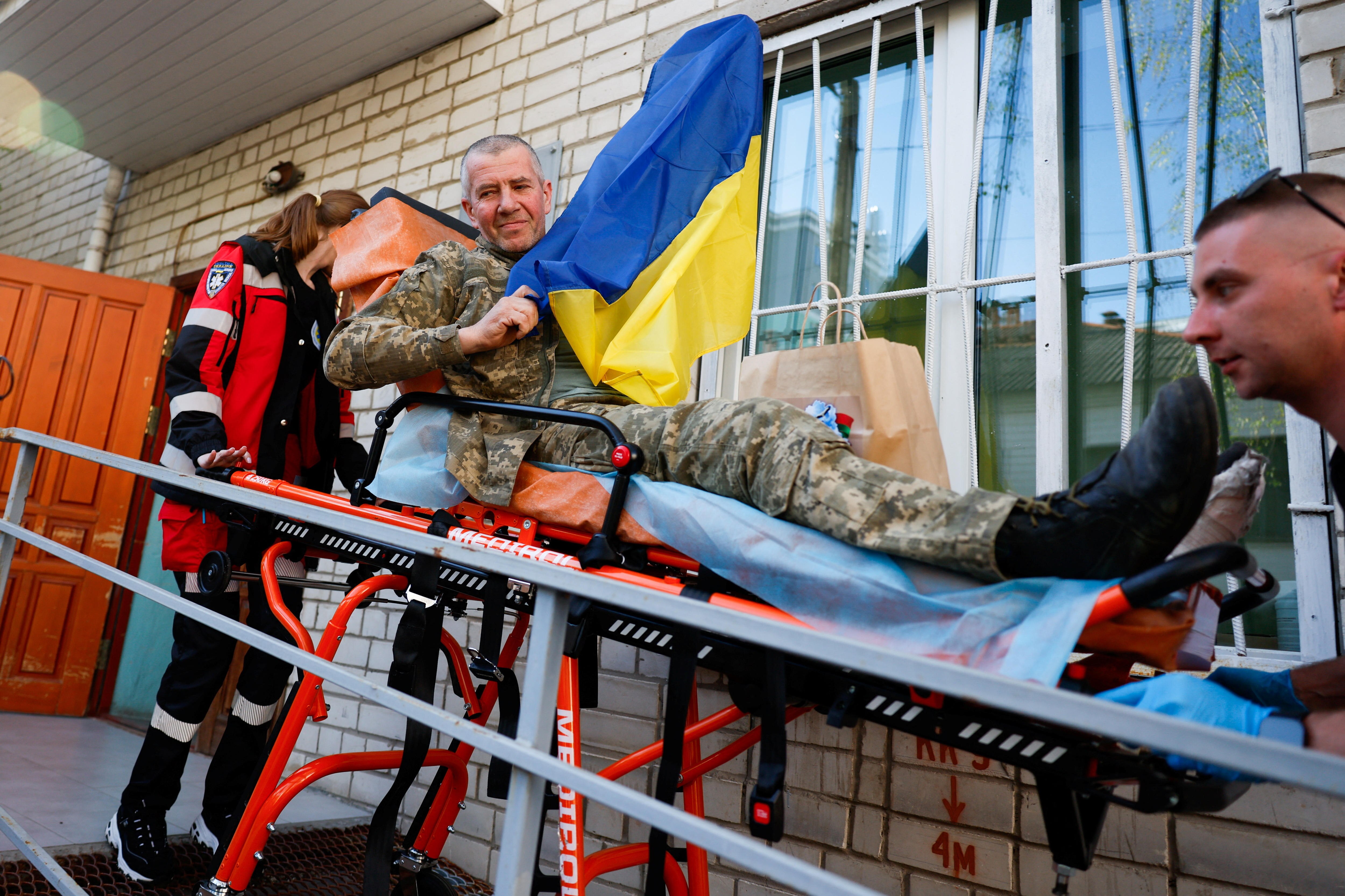 A man in a military uniform sits on a stretcher and holds up a Ukraine flag