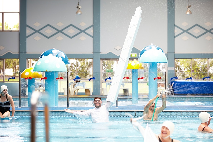 Colour photograph of artist David Capra with 2.5 metre long arm, standing in a pool surrounded by swimmers.