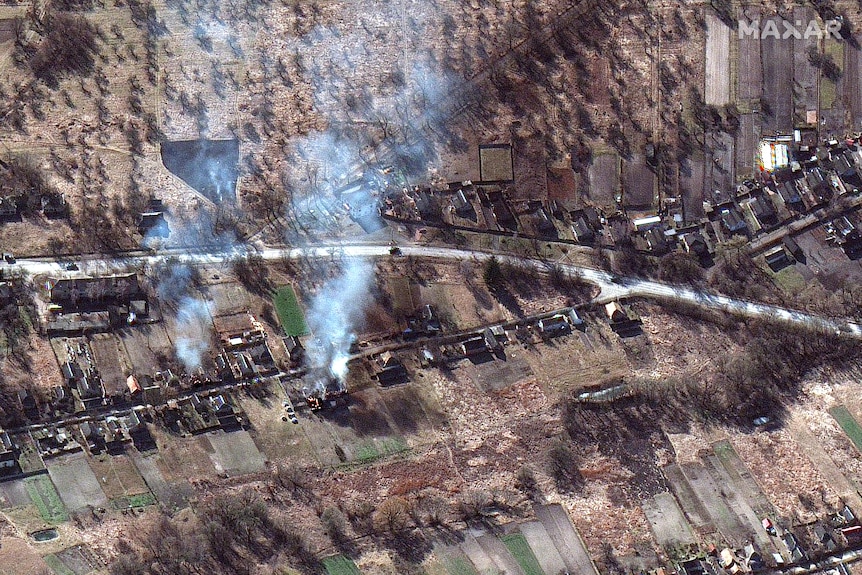 Satellite image showing smoke, burned houses and trees, military vehicles on road. 