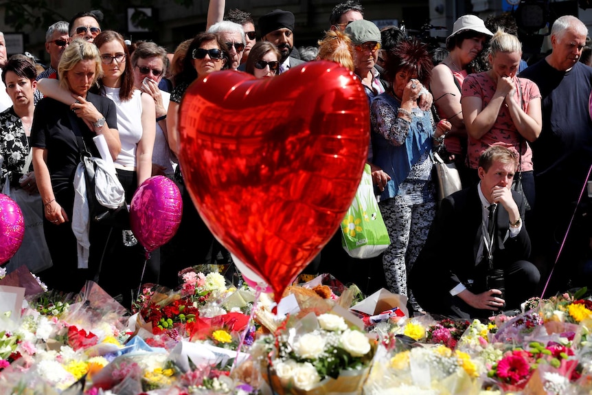 A large group of people stand behind flowers and tributes as a minute's silence is held.