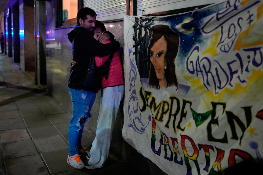 A man and a woman embrace each other on a footpath at night beside a banner featuring a woman's face.