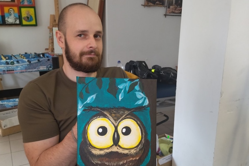 A man with short hair and a beard in a wheelchair holding up a painting of an owl. 