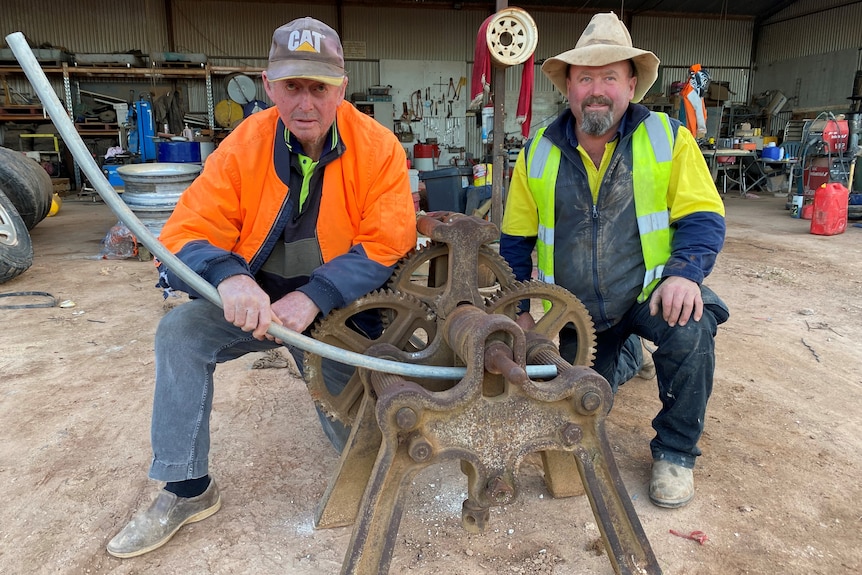 Two men kneeling down behind rusted antique machine with cogs and a metal pole sticking out