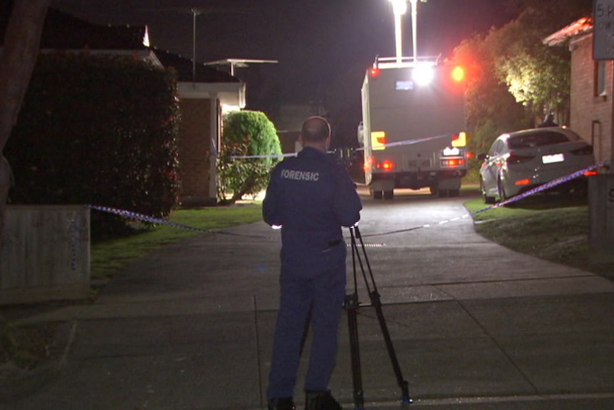A forensic police officer is shown from behind photographing the driveway of a complex of units.