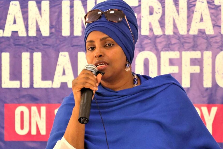 A woman in a blue head wrap speaks into a microphone.