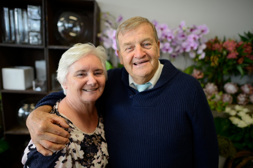 An elderly couple smile with the man's arm around the shoulders