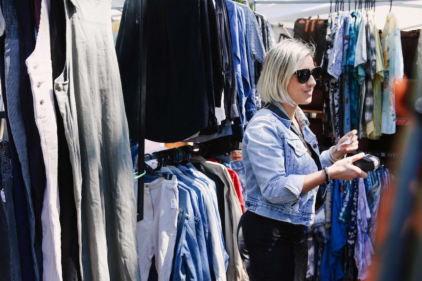 A woman with blonde hair and sunglasses stands among racks of clothes