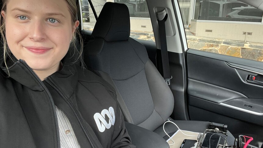 Young woman with brown hair and blue eyes taking a selfie in a stationary car. The Mobile Journalism kit on the seat.