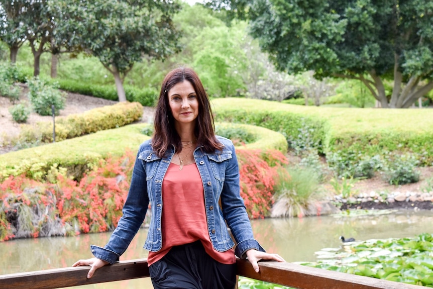 Lea-Ann Marinos standing in the corner of a railing in front of a pond and grassy green park.