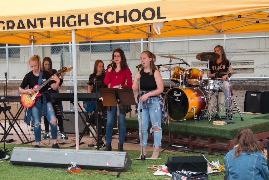 A group of six students singing and playing guitar, keyboard and drums outdoor.