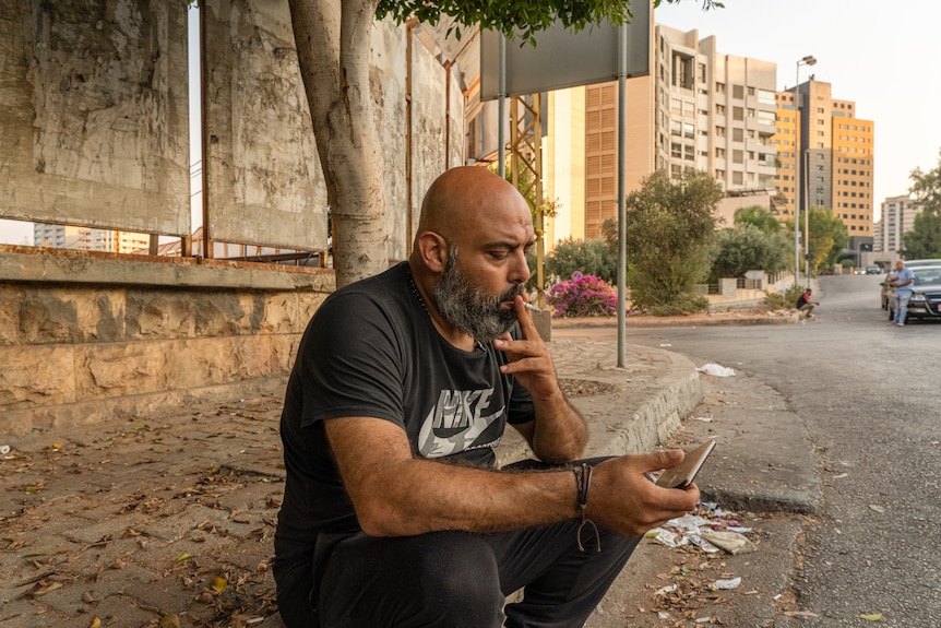 A bald man sits on the side of the road wearing dark clothes and smoking a cigarette.
