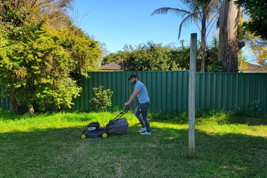 a man wearing a cap, holding a lawnmower as he mows grass in a backyard 