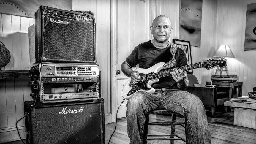 A man sits next to a series of amplifiers and speakers, holding a guitar in his hands.