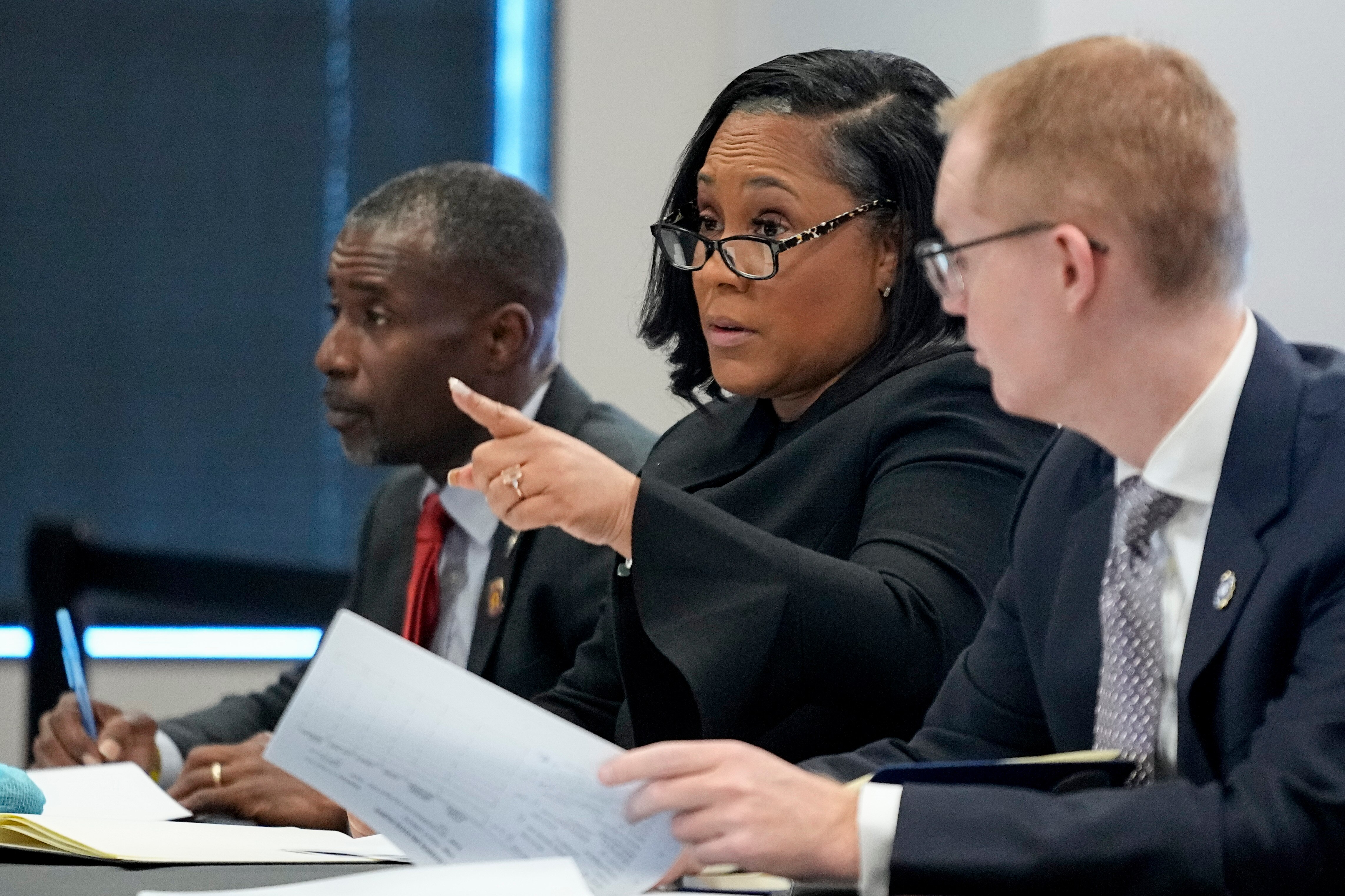 Fani Willis is seated at a desk between two men. She wears black-framed glasses and black clothing. She is pointing.