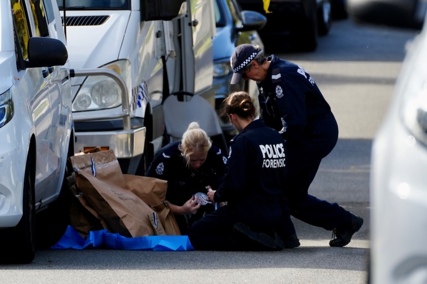 Three police forensic officers hunched over items near a police vehicle.