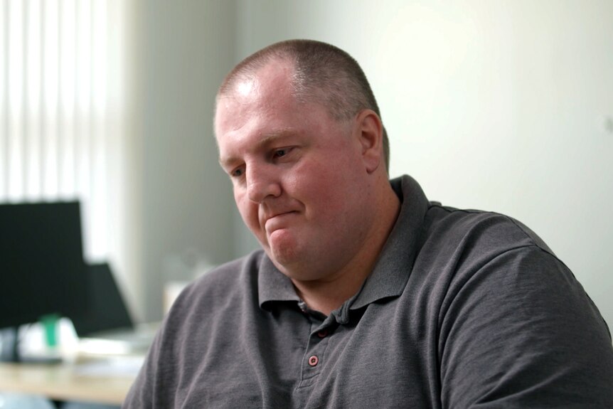 A man sitting in an office with computers behind him looks down, frowning.