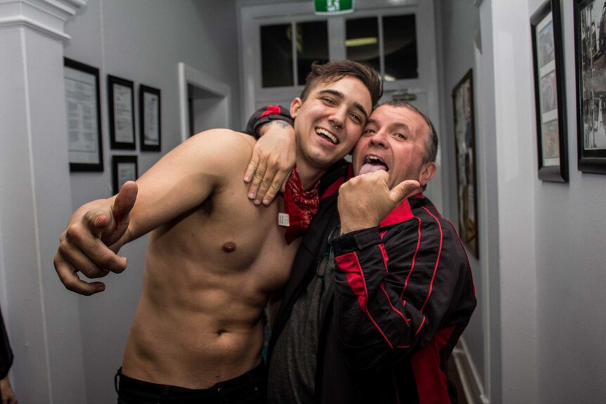 Two men, one shirtless, embrace and smile.
