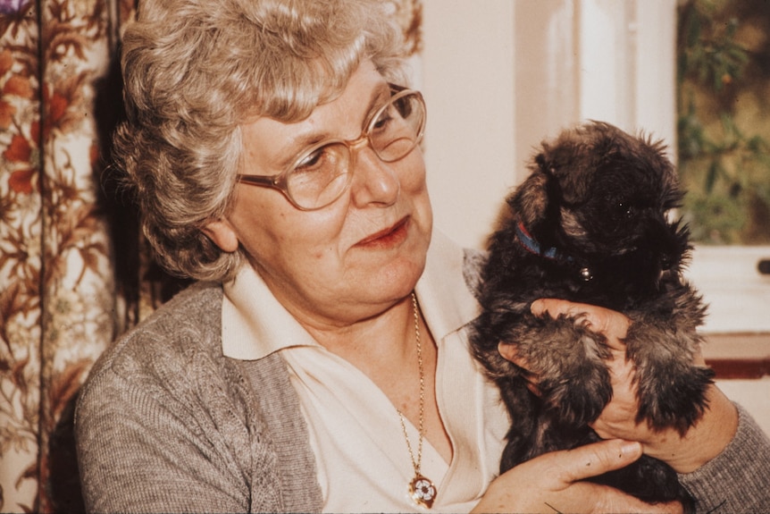 An older woman with glasses and a grey cardigan cuddles a small dog with dark fur