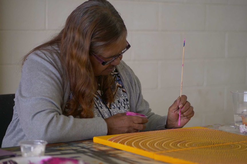 Debra Maher uses a wooden stick to paint pink dots to a yellow canvas, which is neatly covered in dots in a circular pattern.