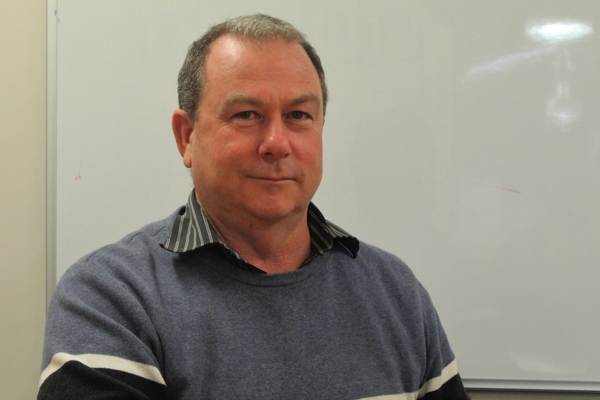 A middle aged man smiles quietly at camera sitting in front of a white board wearing a blue and white striped jumper. 