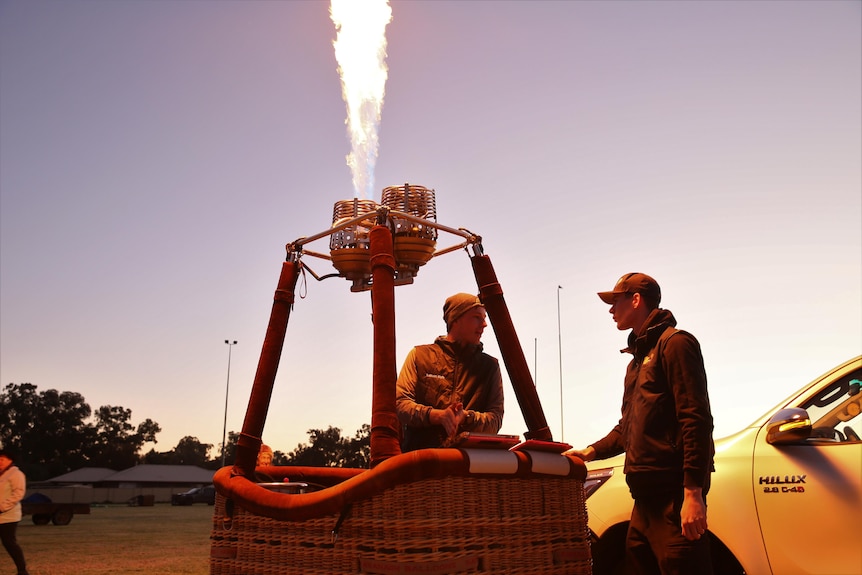 Two men stand at a hot air balloon basket, a burner firing flames up into the dawn sky.