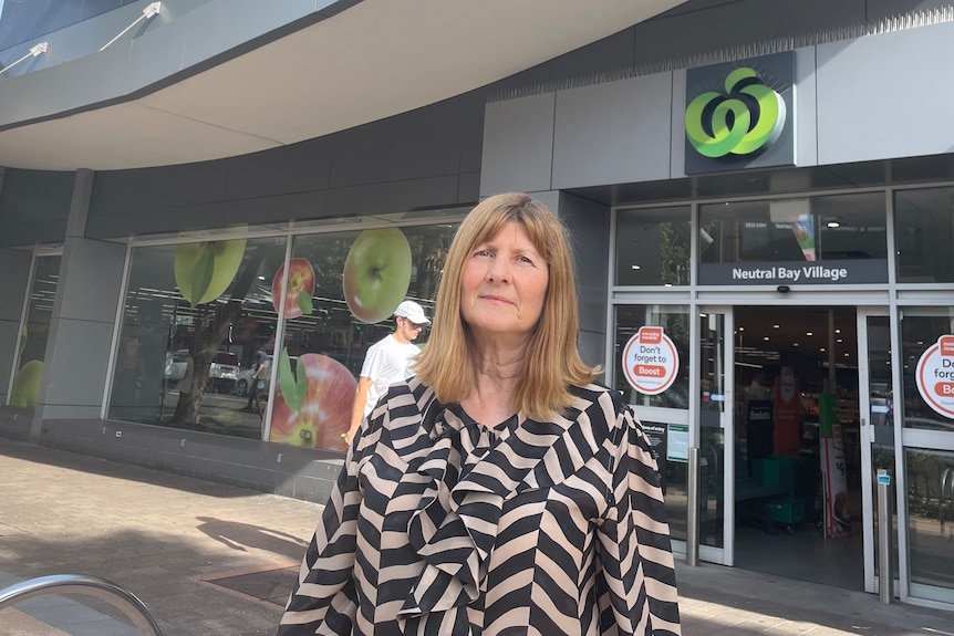 A middle aged woman wearing a beige and black patterned top stands outside a Woolworths supermarket on a clear day.