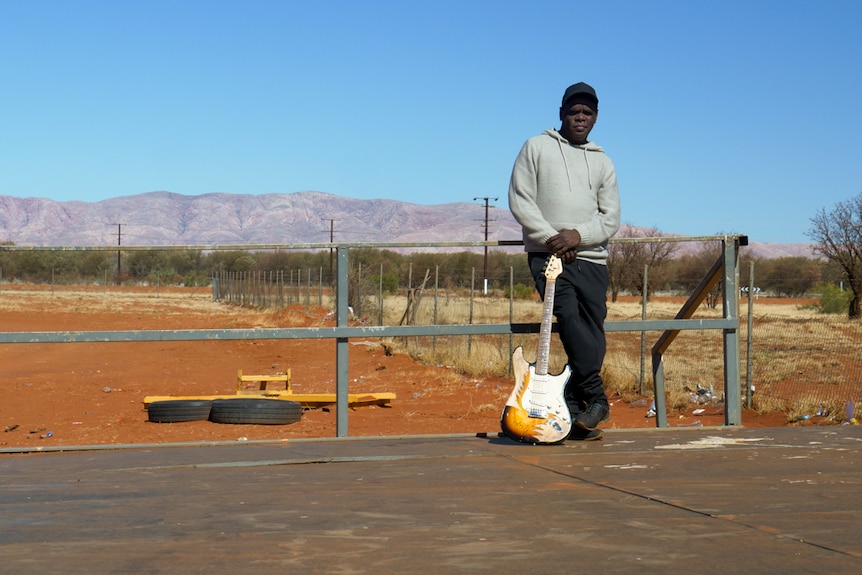 A man in a baseball cap stands next to a guitar, with desert and mountains in the background.