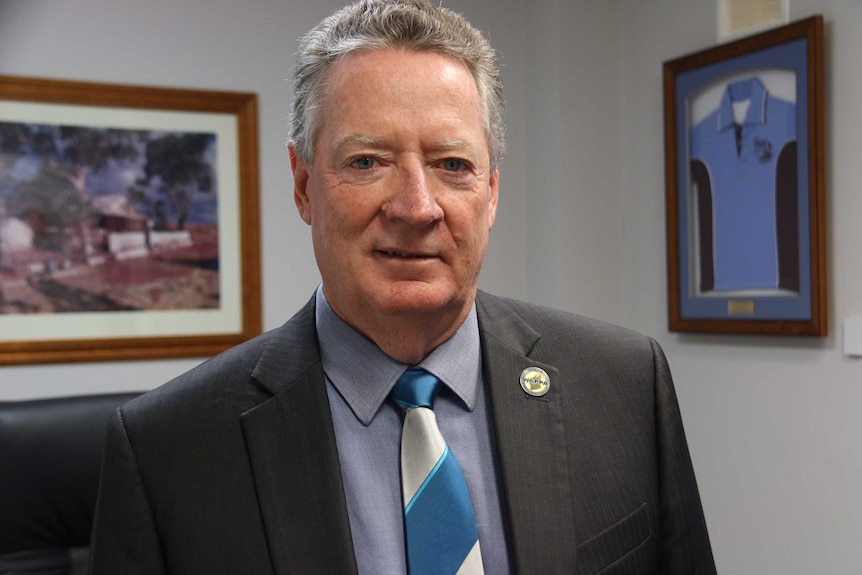 Grey haired man in dark grey suit and two pictures on wall behind him