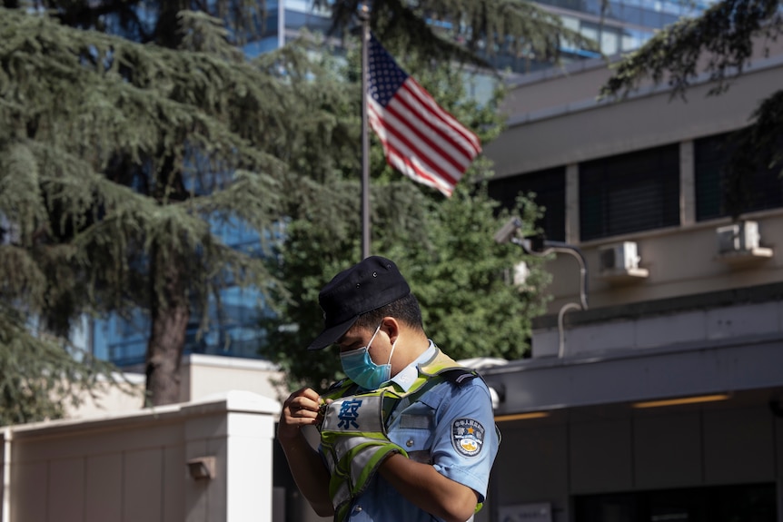 A man in a Chinese police uniform stands in the foreground while behind a wall a US flag stands outside a building